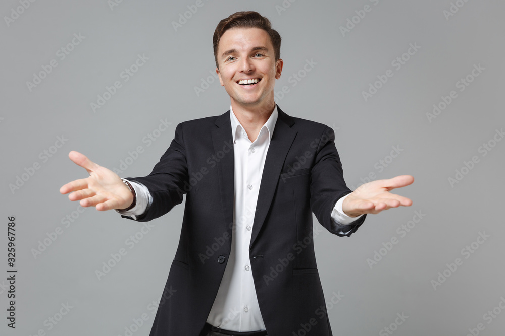Smiling young business man in classic black suit shirt posing isolated on grey background in studio. Achievement career wealth business concept. Mock up copy space. Standing with outstretched hands. Smiling young business man in classic black suit shirt posing isolated on grey background in studio. Achievement career wealth business concept. Mock up copy space. Standing with outstretched hands.
