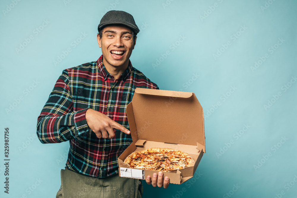 Happy positive young hispanic man from pizza delievery service posing on camera and smile. Point on box with pizza in hand. Stand alone isolated over blue background. Happy positive young hispanic man from pizza delievery service posing on camera and smile. Point on box with pizza in hand. Stand alone isolated over blue background.