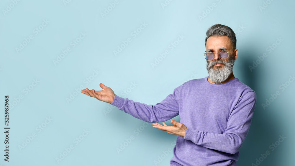 Gray-haired, bearded elderly man in sunglasses, purple sweater. Showing something by hands, posing on blue background. Close up Gray-haired, bearded elderly man in sunglasses, purple sweater. Showing something by hands, posing on blue background. Close up