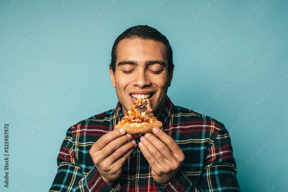 Young joyful hispanic or latin young man biting slice of pizza. Hold eyes closed and enjoy moment. Delisious tasty but unhealthy meal. Guy wear checkered shirt. Isolated over blue background. Young joyful hispanic or latin young man biting slice of pizza. Hold eyes closed and enjoy moment. Delisious tasty but unhealthy meal. Guy wear checkered shirt. Isolated over blue background.