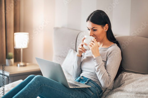 Girl sitting on the bed drinking coffee and using laptop.
