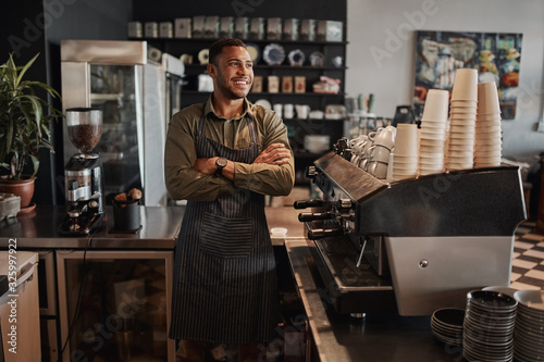 Thoughtful afro-american small coffee shop owner standing behind counter wearing apron with crossed arms looking away photo