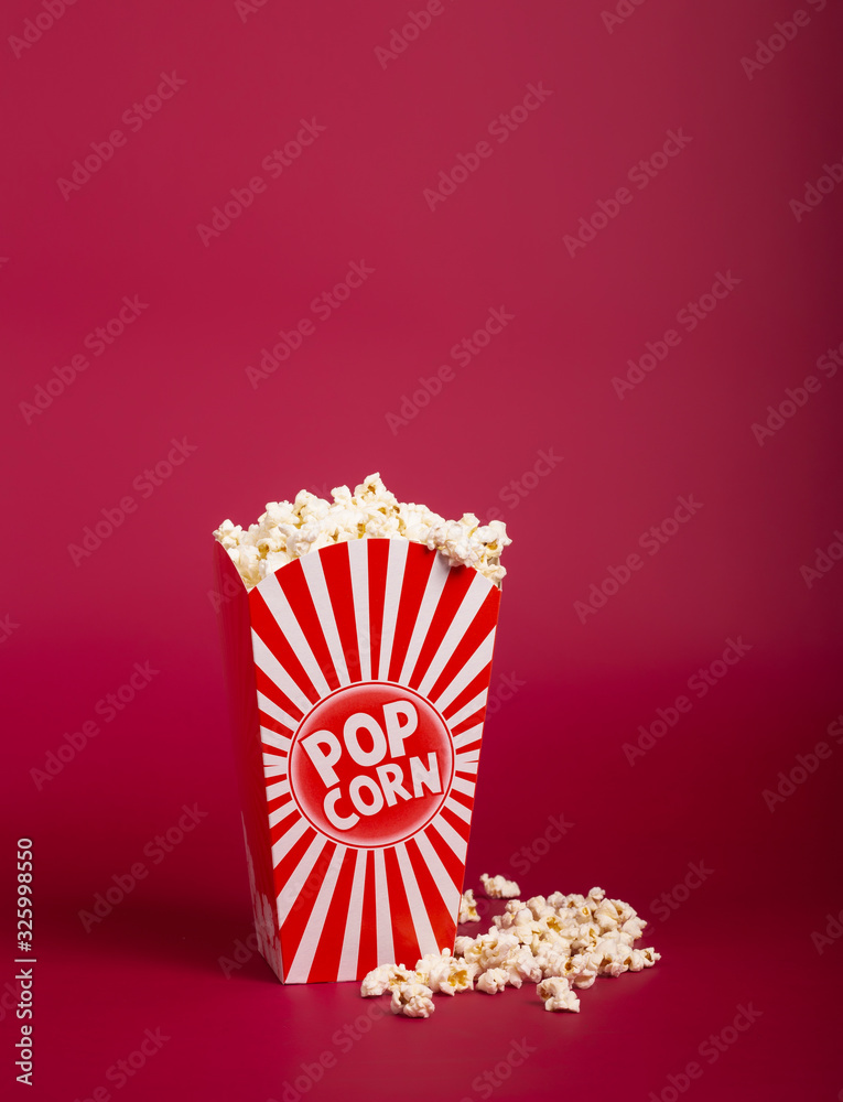 Popcorn in striped bucket on red background. Hot corn scattered from the paper box, copy space. Fast food and movie snack.