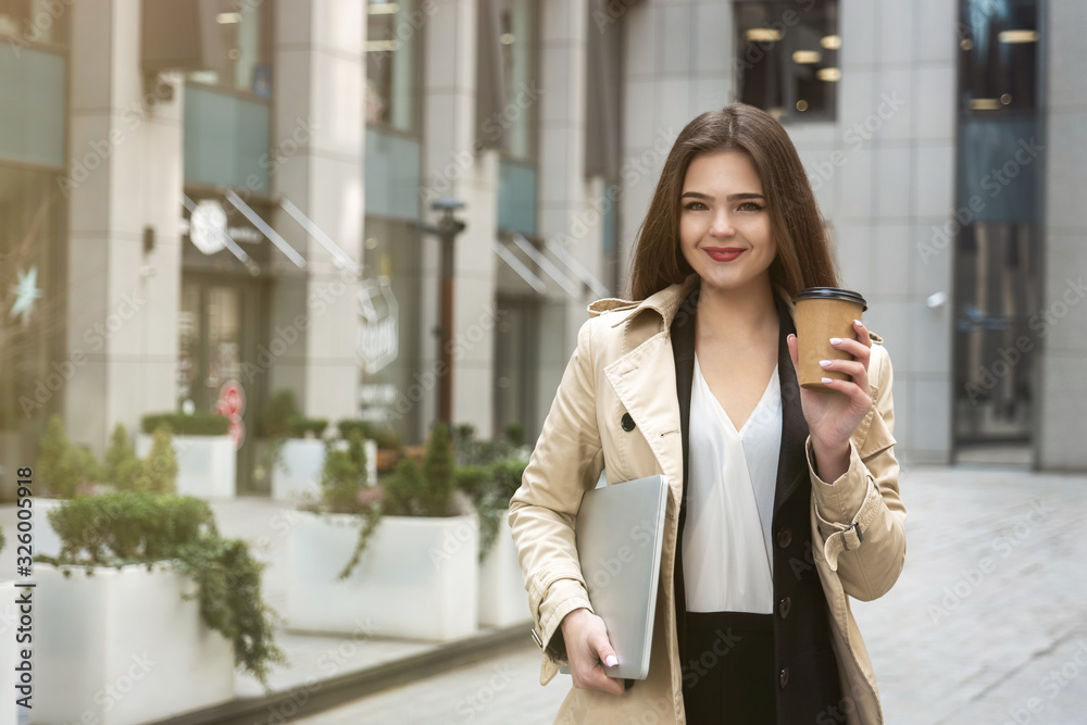 Young business lady holding laptop under arm walking down the street near office center with cup of coffee looking busy, eating on the go concept