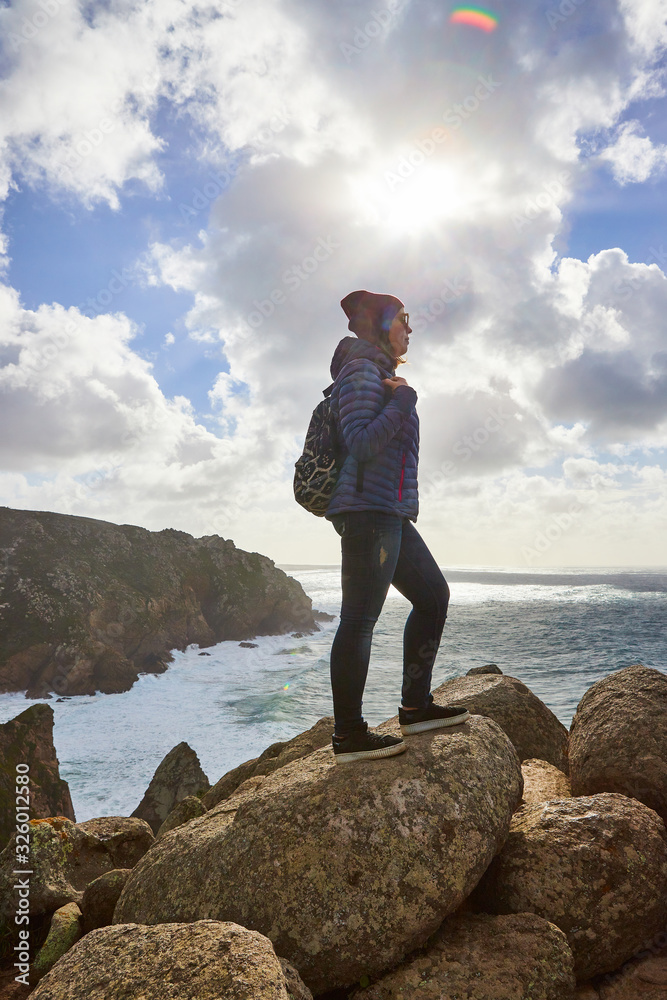 Obraz premium Young woman tourist at Cape Cabo da Roca standing on the rocky edge of the cliff. The waves of the ocean break on the rocks at the bottom of the cliff