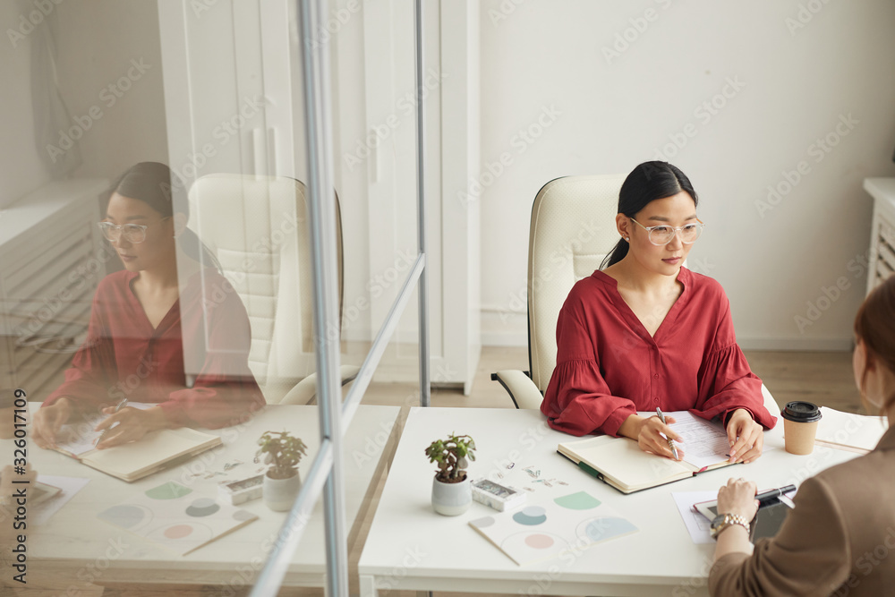 High angle view at modern Asian businesswoman talking to client while working at desk in white office cubicle, copy space