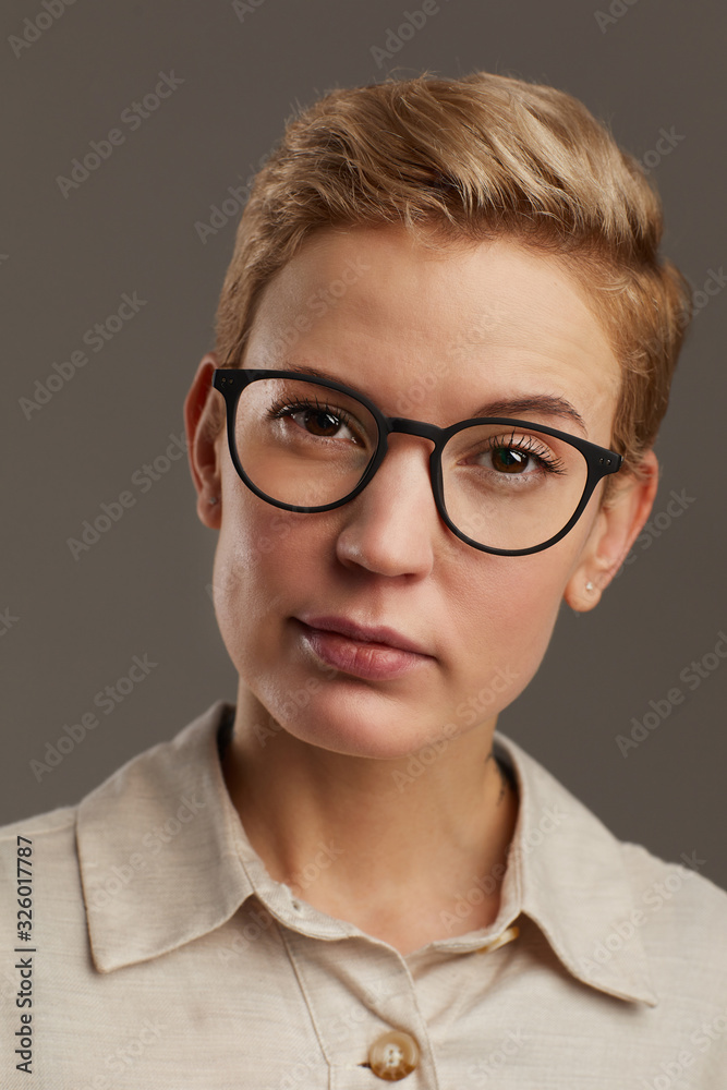 Head and shoulders portrait of modern woman wearing black rimmed glasses with short pixie haircut looking at camera while standing against grey background in studio