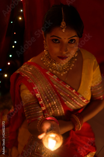 An young and beautiful Indian Bengali woman in Indian traditional dress is sitting while holding Diwali diya/lamp in her hand in front of colorful bokeh lights. Indian lifestyle and Diwali celebration