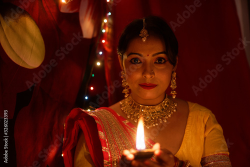 An young and beautiful Indian Bengali woman in Indian traditional dress is sitting while holding Diwali diya/lamp in her hand in front of colorful bokeh lights. Indian lifestyle and Diwali celebration
