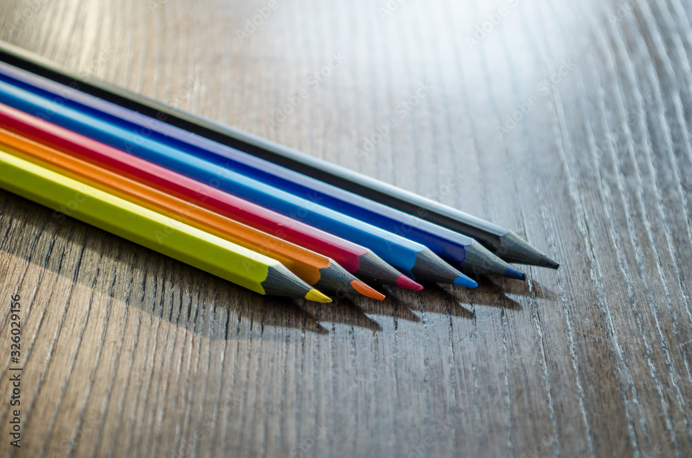 Six colored pencils lie on a dark wooden table in the sunlight