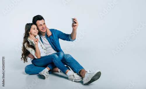 Peaceful photo. Full-length photo of an adult man in denim jacket sitting on the floor with his cute little child and making selfies with her.