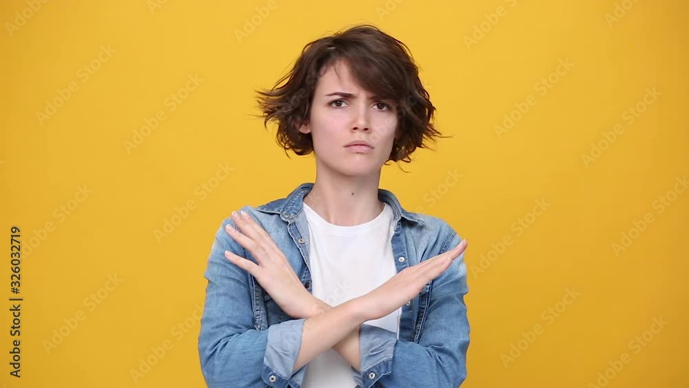 Serious young woman in denim jacket white t-shirt posing isolated on yellow background in studio. People sincere emotions lifestyle concept. Looking at camera say no crossed hands showing stop gesture Serious young woman in denim jacket white t-shirt posing isolated on yellow background in studio. People sincere emotions lifestyle concept. Looking at camera say no crossed hands showing stop gesture