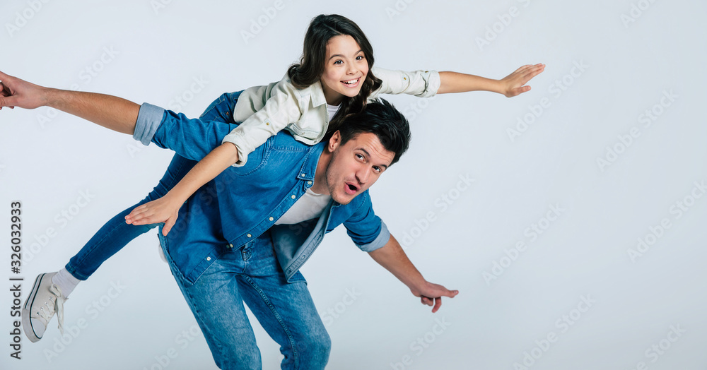 Enjoying the flight. Cheerful girl with long hair is riding on her father’s back, while they are both having fun, laughing and smiling together. Enjoying the flight. Cheerful girl with long hair is riding on her father’s back, while they are both having fun, laughing and smiling together.