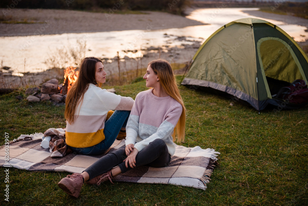 Two women near tent and campfire. Friends are sitting on the grass on a blanket and talking. Enjoy the scenery of mountains and rivers. Hiking, vacation and friendship concept