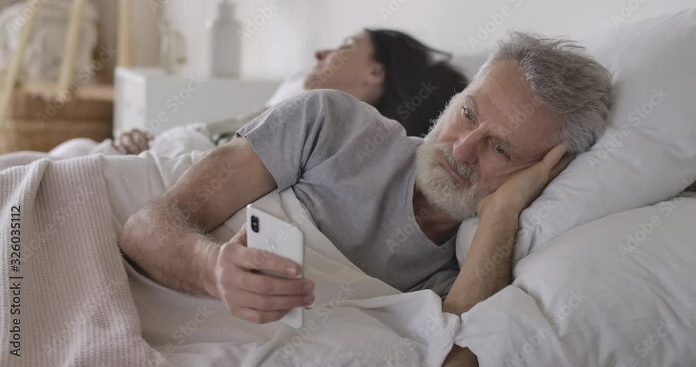 Close-up portrait of senior Caucasian man lying in bed and using smartphone as his wife sleeping at the background. Handsome mature man using modern technologies. Leisure, morning. Cinema 4k ProRes HQ Close-up portrait of senior Caucasian man lying in bed and using smartphone as his wife sleeping at the background. Handsome mature man using modern technologies. Leisure, morning. Cinema 4k ProRes HQ