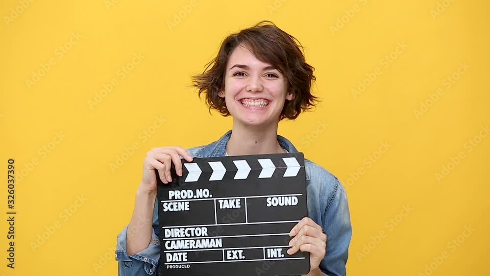 Smiling young woman in denim jacket white t-shirt posing isolated on yellow background in studio. People sincere emotions lifestyle concept. Look at camera hold classic black film making clapperboard