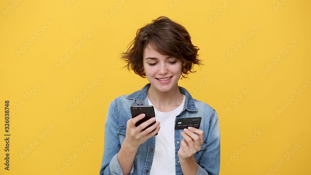 Smiling woman in denim jacket white t-shirt posing isolated on yellow background in studio. People sincere emotions, lifestyle concept. Looking using mobile phone hold credit bank card online shopping Smiling woman in denim jacket white t-shirt posing isolated on yellow background in studio. People sincere emotions, lifestyle concept. Looking using mobile phone hold credit bank card online shopping