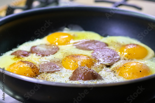 Fried eggs with sausage and seasonings in a black pan on a gas stove. Tasty and nutritious breakfast for the family. Selective focus. Closeup view