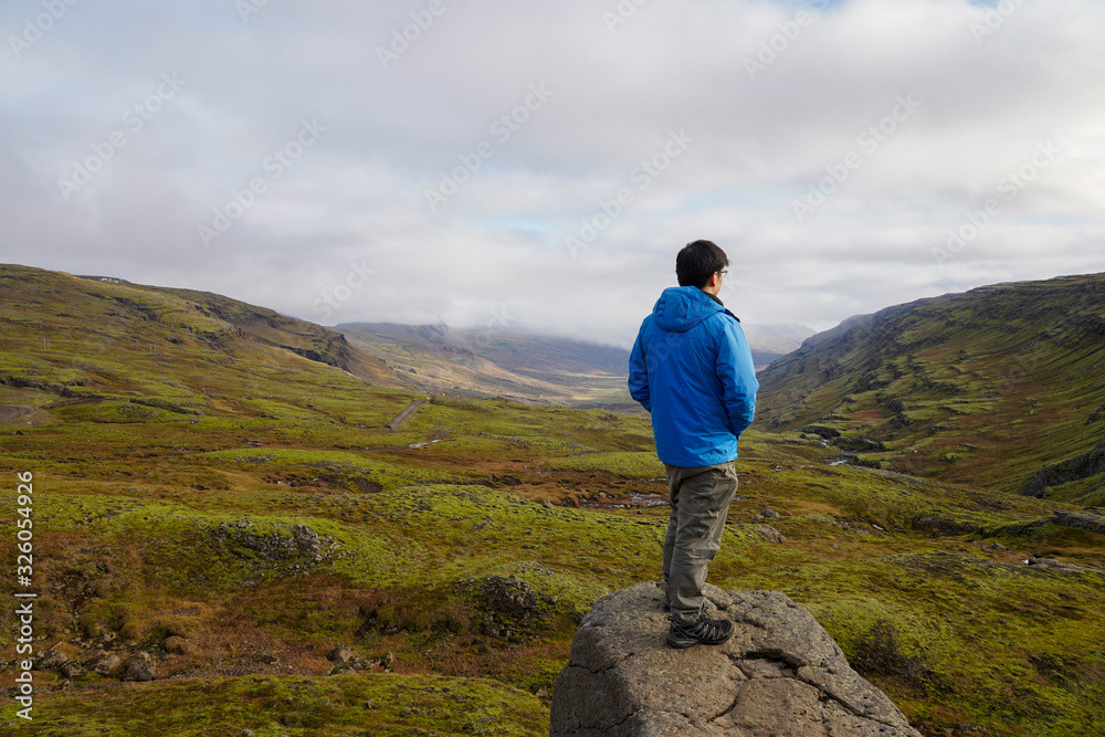 young asian man standing pose on the rock look out to mountain terrace view with grass field, Iceland horizontal image