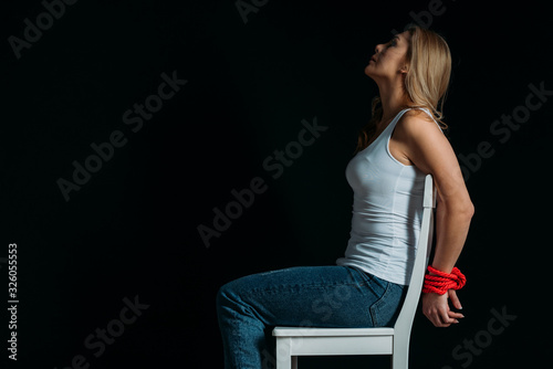 Side view of woman with tied hands sitting on white chair isolated on black