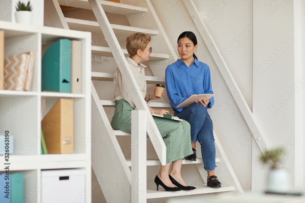 Full length portrait of two modern businesswomen sitting on stairs in office and chatting, copy space Full length portrait of two modern businesswomen sitting on stairs in office and chatting, copy space