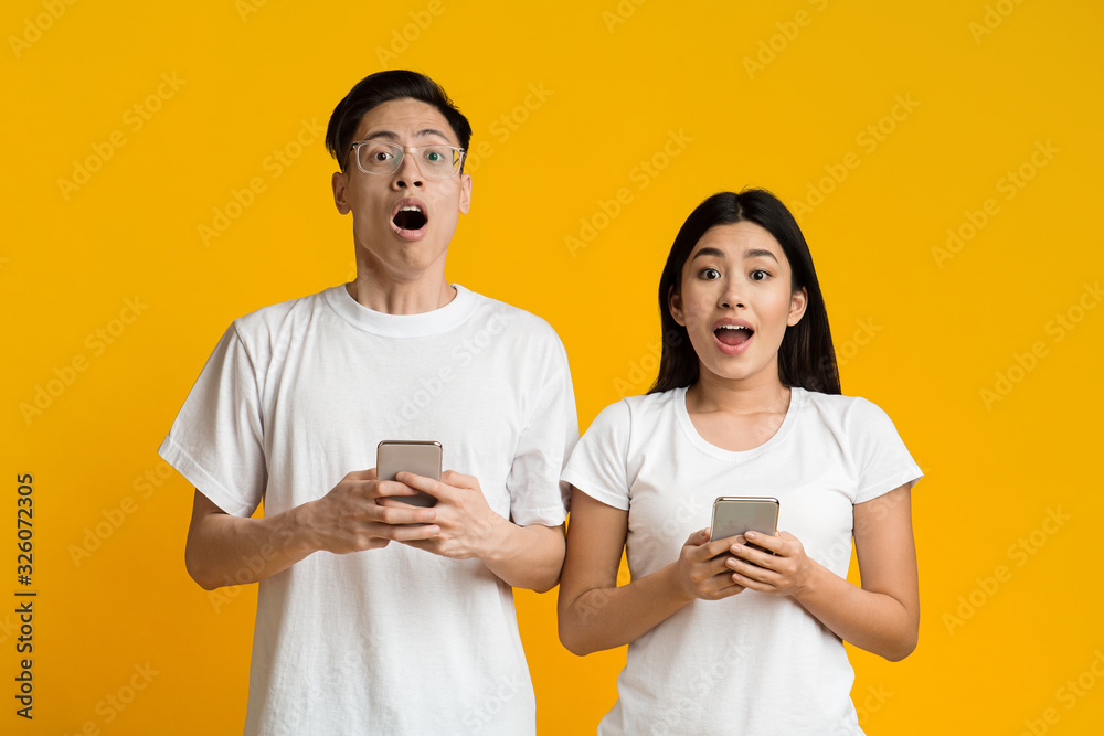 Amazed asian couple holding smartphones over yellow background Amazed asian couple holding smartphones over yellow background