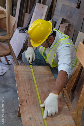 Handsome man wearing safety vest and yellow helmet,measuring timber board,for wood work design,at factory