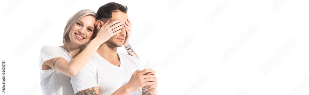 smiling young tattooed girl covering boyfriend eyes while man using smartphone isolated on white, panoramic shot