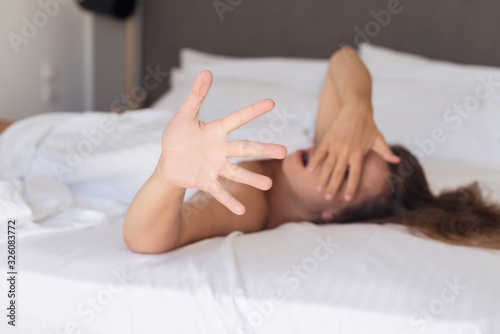 Young woman hides her surprised and indignant face from the camera lying on a bed in the bedroom and covered sheet
