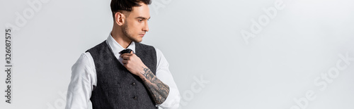 handsome tattooed elegant bridegroom adjusting tie isolated on grey, panoramic shot
