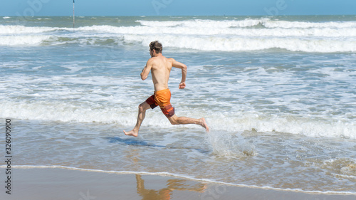 Handsome man with athletic body running around the beach line near the sea.Fit runner on the beach with sea waves background. Beach activities concept.