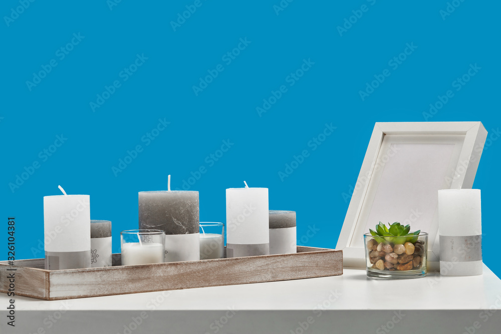 White table with green succulent in glass pot, some different sized candles in a wooden stand and empty photo frame. Blue background. Close up White table with green succulent in glass pot, some different sized candles in a wooden stand and empty photo frame. Blue background. Close up