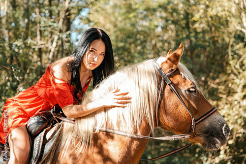 A young beautiful brunette woman in a red dress sits astride a brown horse. Summer. Sunlight