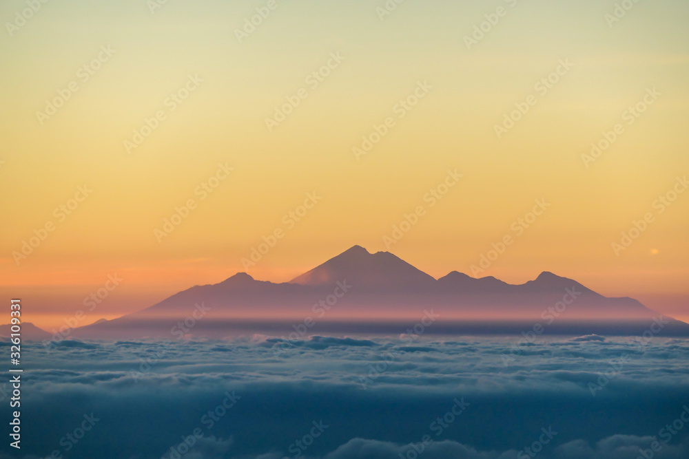 A distant view on Mount Rinjani on Lombok, Indonesia, captured from Mount Batur on Bali, during the morning golden hour. There is a slight fog around the lower parts. Mysterious and magical sunrise.