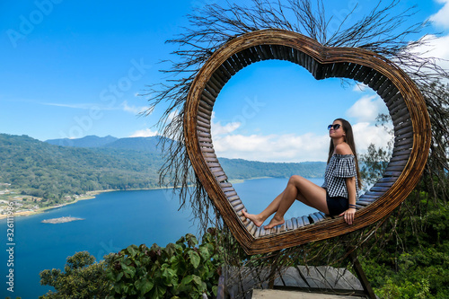 Woman with long hair enjoying her time on the wooden heart shaped seat with The Twin Lake in the background, Bali, Indonesia. The seat is high above the lake. She is having fun. Touristic attraction