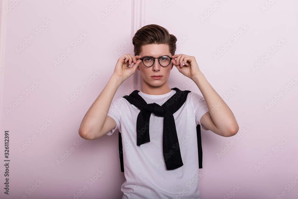 Fototapeta premium Model of a stylish handsome young hipster man stands and straightens fashionable glasses near a pink wall indoors. Attractive guy in classic black-white clothes with trendy hairstyle posing in studio.