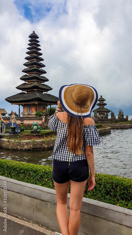 A woman in shorts and straw hat standing in front of main building of Ulun Danu Temple, Bali, Indonesia. The girl is admiring the beauty of Hindu water temple. Sacred place. Discovering new cultures.