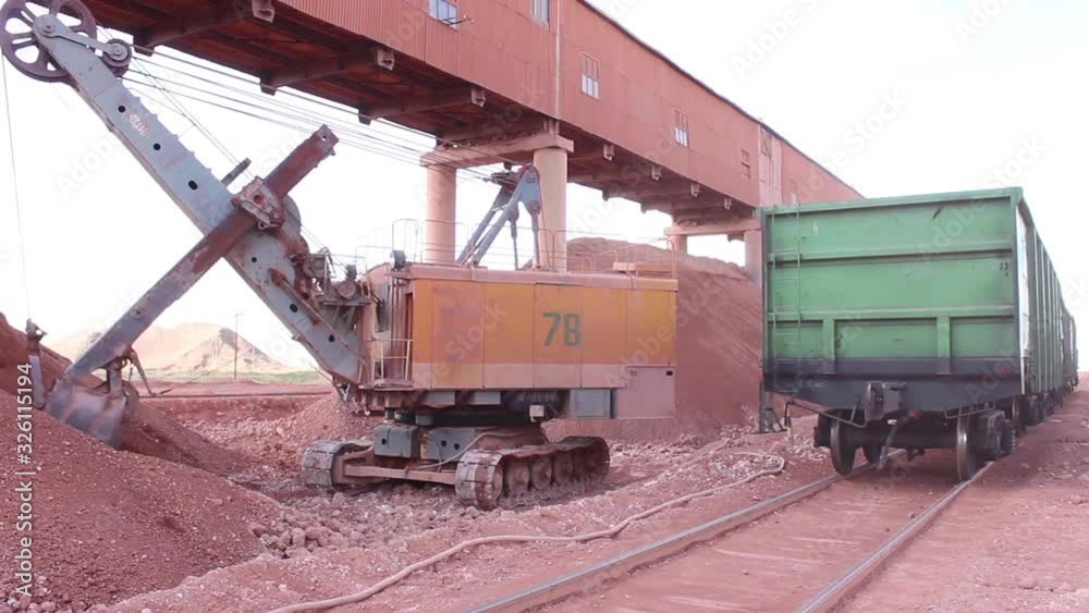 A large quarry excavator loads a railway car with minerals (red soil ). The excavator does its hard work. A large quarry excavator loads a railway car with minerals (red soil ). The excavator does its hard work.