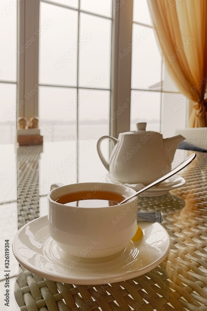 One hot tea white cup on rattan table in hotel restaurant. Tea with lemon for breakfast at a cafe in the resort. Morning tea cup with teapot on table in country house One hot tea white cup on rattan table in hotel restaurant. Tea with lemon for breakfast at a cafe in the resort. Morning tea cup with teapot on table in country house