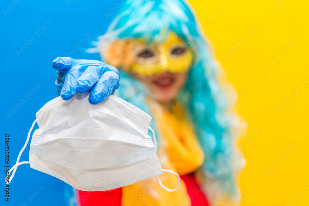 Fototapeta premium Concept of protection against coronavirus at carnival. A bright woman in a blue wig, yellow carnival mask, and medical gloves. A girl in a festive costume holds a health mask from Virus crown in hand