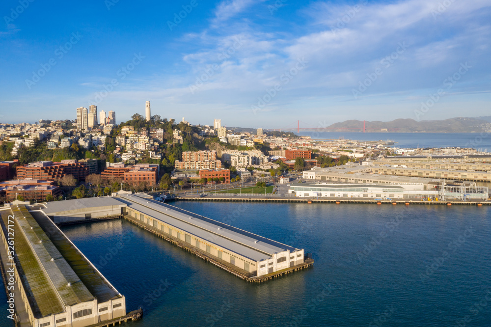 San Francisco, California, daytime, sunrise, aerial view of North Beach with Coit Tower and Golden Gate Bridge visible. Blue sky with clouds and golden light. Embarcadero area in foreground. 