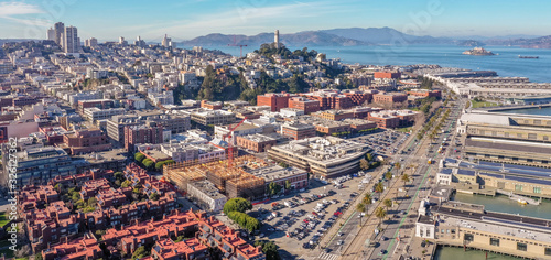 San Francisco, California, daytime, sunrise, aerial view of North Beach with Coit Tower and Golden Gate Bridge visible. Blue sky with clouds and golden light. Embarcadero area in foreground. 