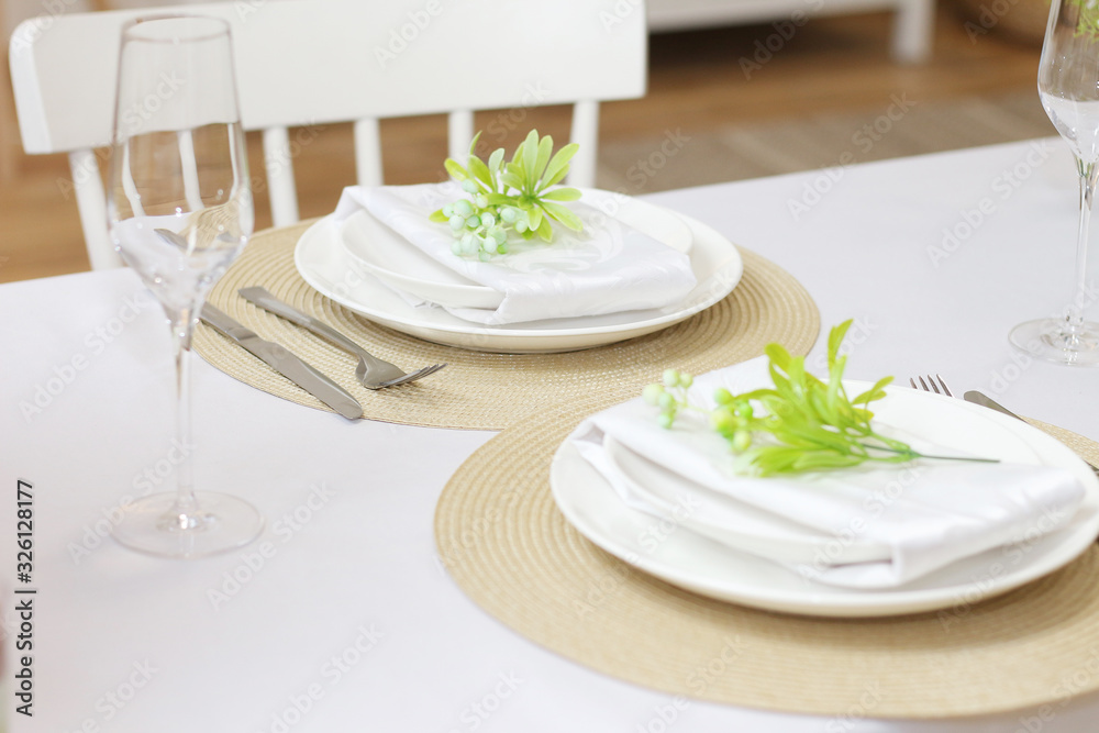 .Plates with twigs of artificial flowers on beige round napkins, glasses and cutlery on a white table..Festive table setting for two persons..
