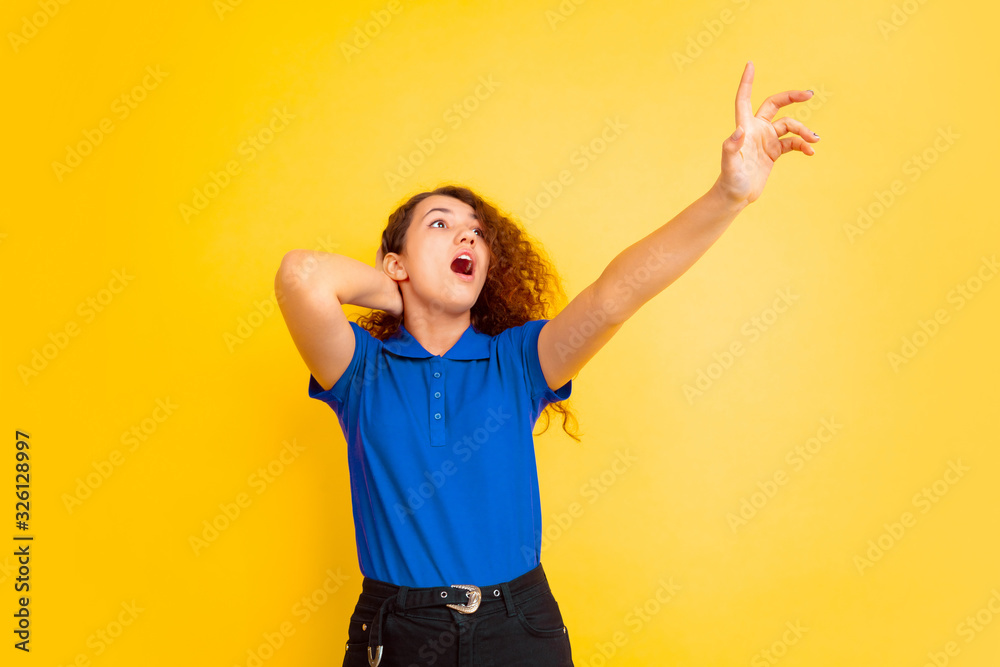 Astonished pointing up. Caucasian teen's girl portrait on yellow studio background. Beautiful female curly model in shirt. Concept of human emotions, facial expression, sales, ad, education. Copyspace Astonished pointing up. Caucasian teen's girl portrait on yellow studio background. Beautiful female curly model in shirt. Concept of human emotions, facial expression, sales, ad, education. Copyspace