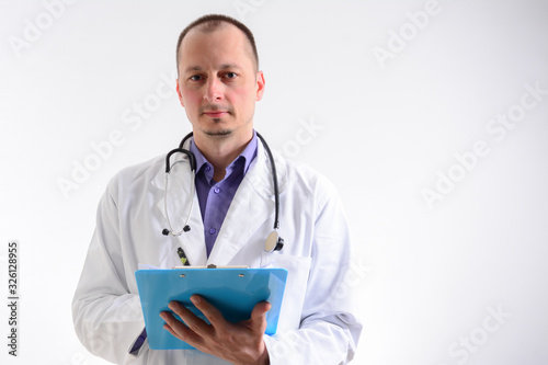 Male doctor holding clipboard and looking at camera while standing against white background. Caucasian male doctor holding blank paper.