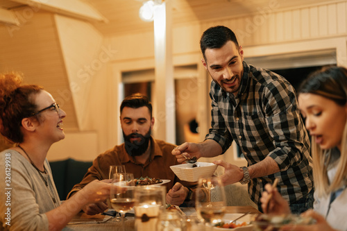 Happy man having fun with friends while serving them food at dining table.