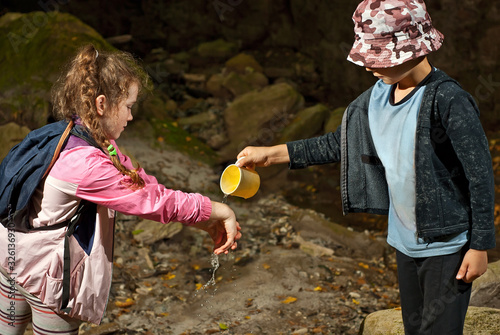 Girl in a pink jacket washes her hands. A boy pours water from a mug near a spring. Children help each other to wash their hands. Concept of assistance and mutual support.