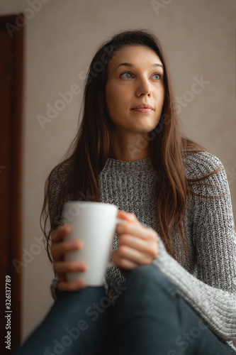 Beautiful girl holding a Cup of coffee or tea, sitting on the bed near the window.