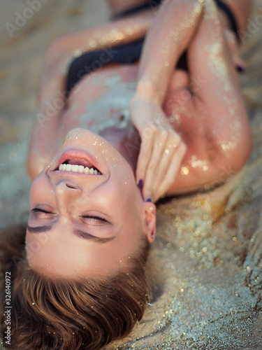 Beautiful tanned woman in a black bikini lies on the sand sunbathes. Sensual girl luxurious gorgeous trendy covered with gold sparkles. Shine attractive sexy. Sea vacation spa paradise sunblock beach