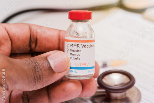 measles mumps and rubella vaccine concept african american doctor holding syringe in front of vaccination vial and stethoscope and clipboard with checklist photo